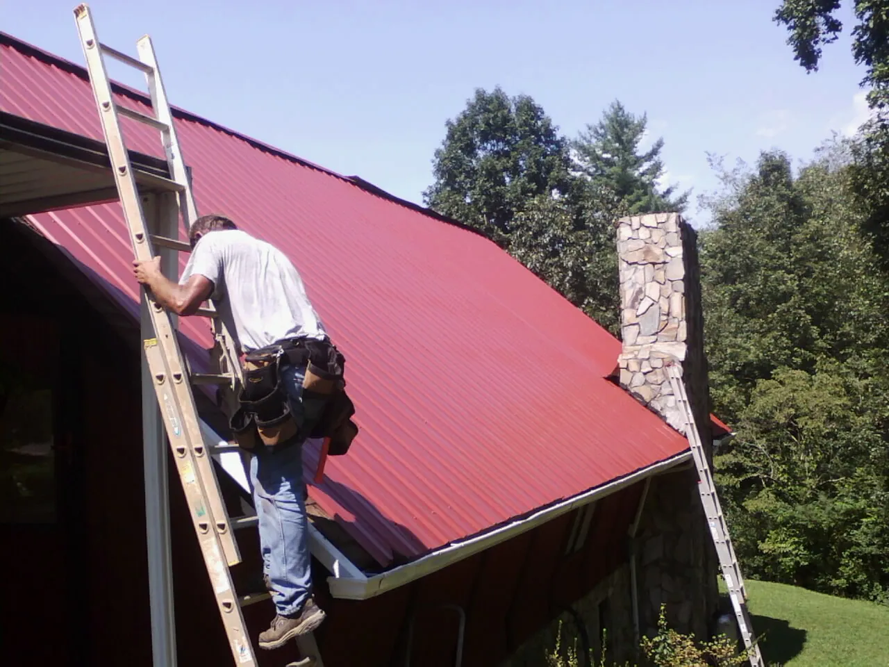 Roofer on a ladder beside a red metal roof and stone chimney — Soffit & Fascia Repair by Roofing Brothers in Cardwell